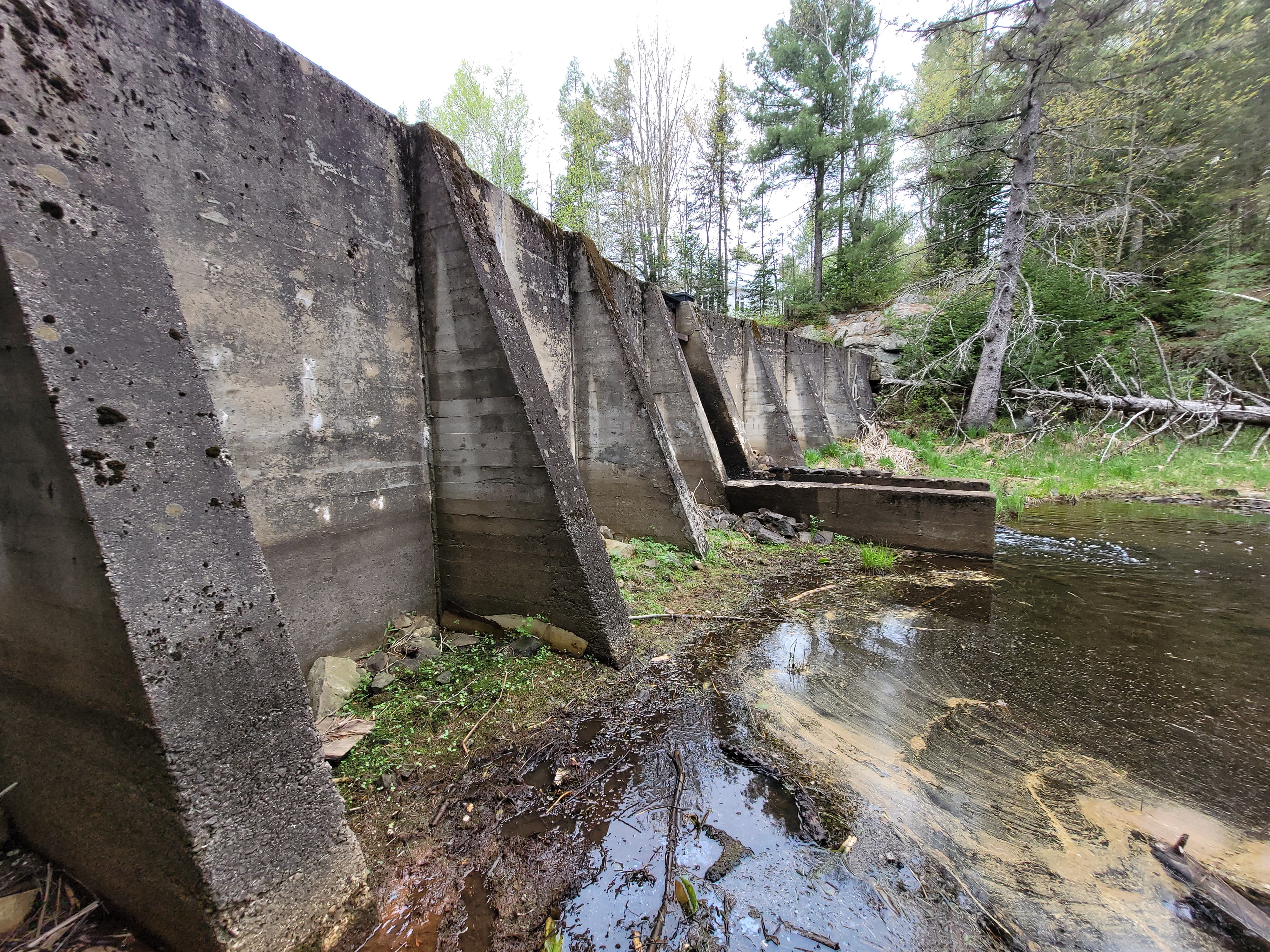 Mur en béton d’un barrage ancien visible en bord de cours d’eau, intégré dans un environnement forestier au Québec